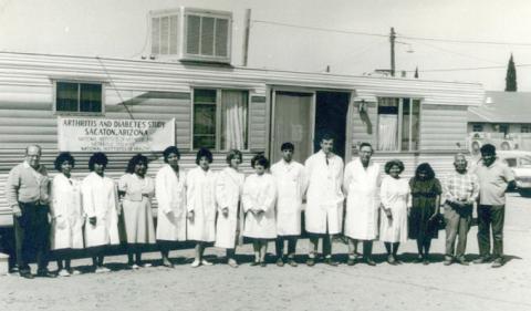 A black-and-white photo shows a group standing in front of a trailer.