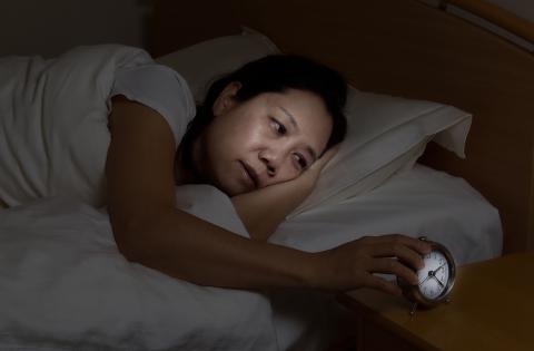 A woman lies awake in bed looking sleepily at alarm clock on nightstand.