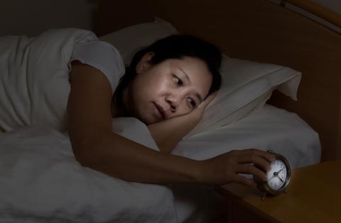 A woman lies awake in bed looking sleepily at alarm clock on nightstand.