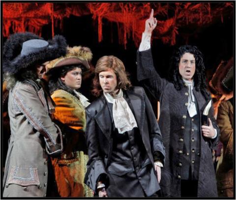 Austin in coattails and a long, curly-haired wig points a finger to the sky as he sings with 3 others in the troupe..