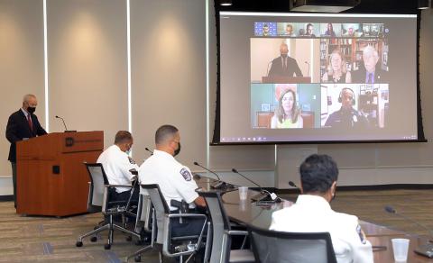Male speaker at podium in conference room with uniformed officers seated around table looking at projector screen