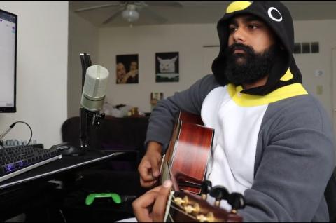 Shaikh, dressed in a hoodie, sits in his room strumming his guitar in front of a microphone and keyboard.