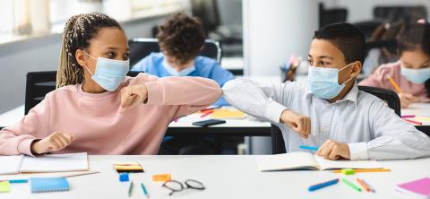 Two children elbow bump in a classroom, with pens, paper and other school supplies on desk in front of them.