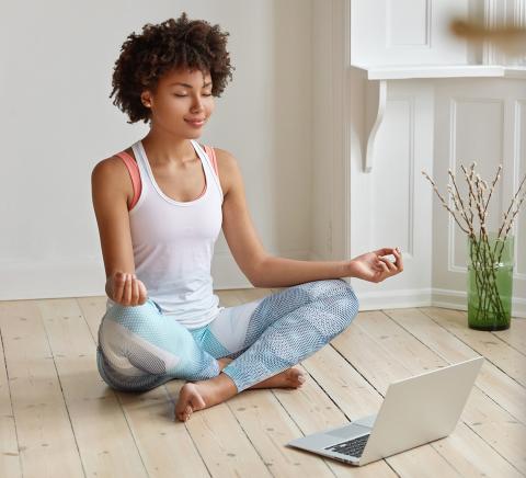 A young Black woman sits on the floor cross-legged, with palms up, fingertips touching, meditating in front of her laptop.