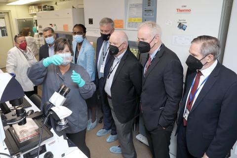 Woman in mask holds up glass flasks in gloved hands as masked people look on