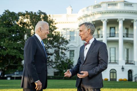 Side view of President Joe Biden and Bill Nye standing on the lawn talking, White House in the background