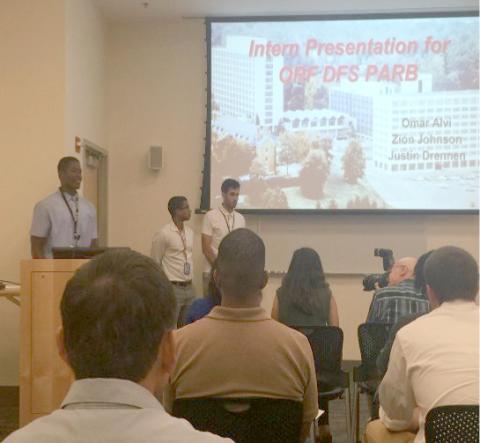 Audience view of Johnson at podium with other interns beside him and slide projected on screen in background.