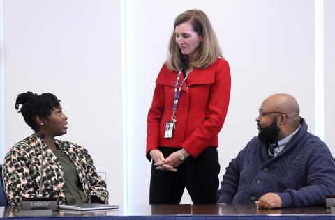 A woman standing talks to a woman and a man seated at a conference table.