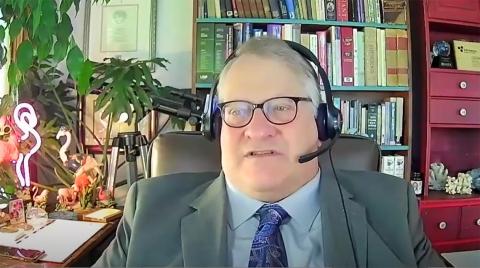Dr. Betz speaks into headset seated by desk in front of a bookcase and red hutch