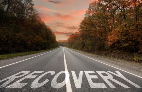 A highway lined with trees and a peachy sky at sunset up ahead; painted on the road is the word "recovery"