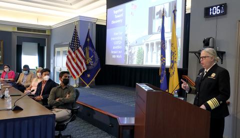 Levine speaks at an NIH podium, with event title slide projected on screen beside her