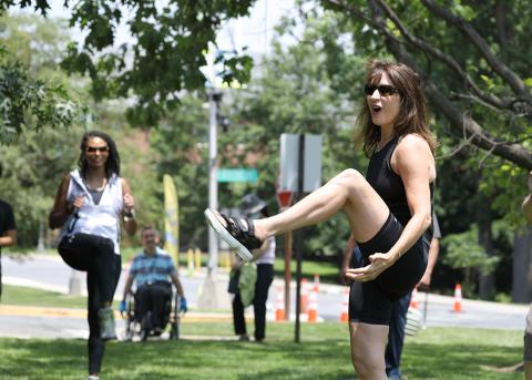 Fitness instructor does a knee lift as a woman follows and man in wheelchair looks on; orange cones behind mark route