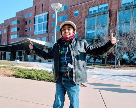 Caesar stands smiling with arms extended and both thumbs up in front of the Clinical Center during a recent checkup.