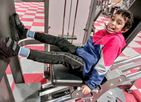 A smiling Caesar sits with his feet up on a leg press machine at the gym