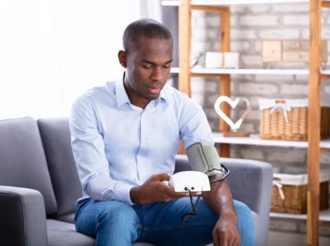 An African American man with cuff around his arm, holding blood pressure machine, sitting on couch in his home