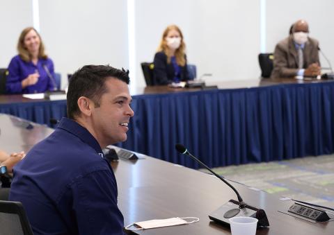 A profile of Greenberg seated, with McGowan, Schwetz and Johnson seated at table, draped in blue cloth, listening.