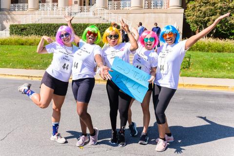 Five women wearing wigs and sunshades in dayglo colors of the rainbow, strike humorous poses.