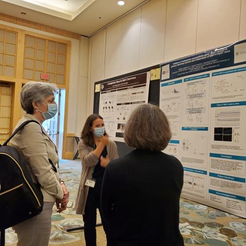 Majocha, in face mask, shown signing in front of her poster as visitor observes with interpreter's back to camera.