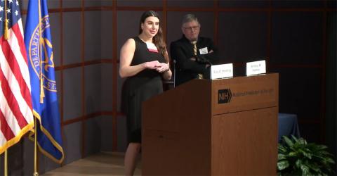Holmes stands at podium, hands clasped, speaking into microphone as Harding stands beside her.