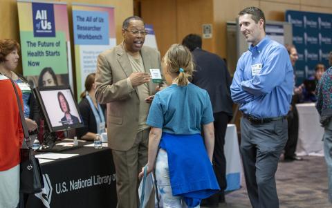 Three attendees stand chatting by All of Us information table in the hallway in Natcher.