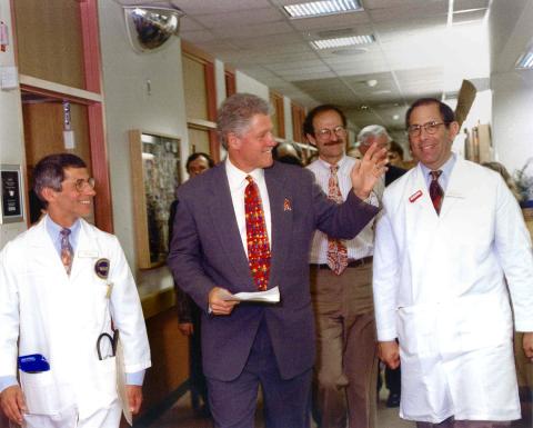 Clinton smiles and waves as he walks through a hospital corridor flanked by Fauci and Gallin