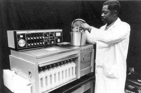 B&amp;W image of Black man standing at a machine with a silver cylinder on top
