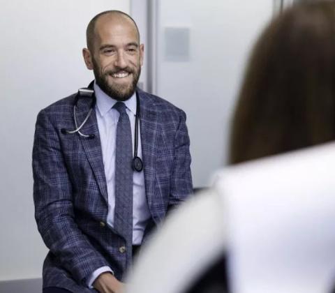 A smiling Diamond, wearing a suit and stethoscope, sits with a patient.