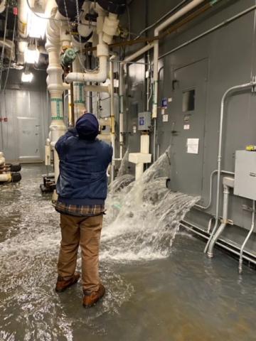 Man with back to camera adjusts device on pipe as water gushes from wall and door in front of him.