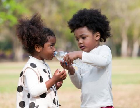 Two children in the park, the older one holding a water bottle to the younger child's mouth