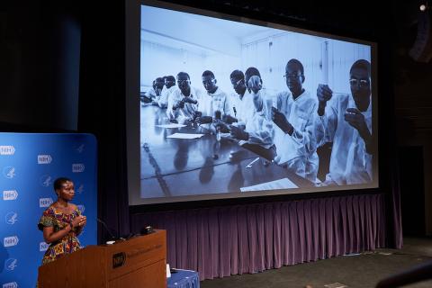 Dr. Sey speaks at podium with slide of scientists at conference table in Ghana on screen behind her.