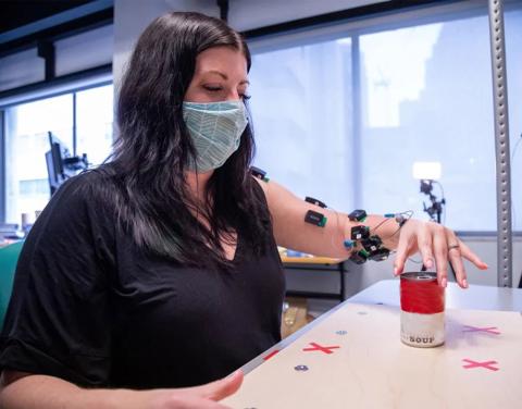 A woman sits with a medical device strapped to her left arm, her left hand grasping and moving a can.