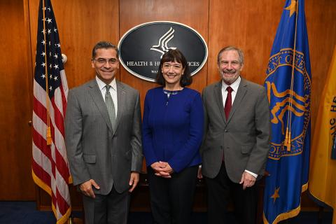 Becerra on left with Bertagnolli in blue shirt and Lowy in front of U.S. and HHS flags