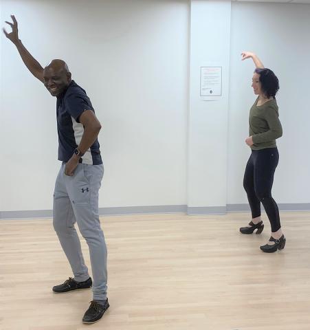 Bacanamwo and Burgess dance with one hand on hip, the other arm high in the air, in the gym