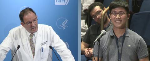 Holland talks into mic at podium; on right, a man stands at mic asking question next to seated audience