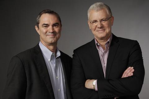 Smith and Johnson smile shoulder-to-shoulder. Smith, left, wears a black suit jacket and blue shirt, and Johnson wears glasses, a black suit jacket and checkered red and white shirt.