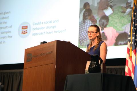 Woman speaks at podium in front of slide show presentation