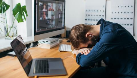 Stock photo of a man with his head on the desk, a Zoom meeting on the 2 screens on his desk