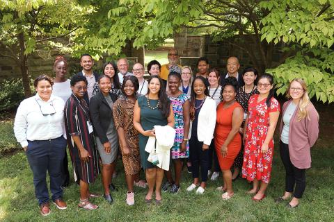 A large group poses together on a grassy area beneath a tree with a brick building in the background.