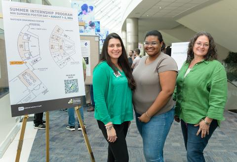 Three women stand smiling in the hall of Natcher Conference Center next to a summer internship day poster.
