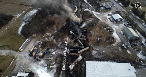 looking down at train cars tilted and off the tracks, large plume of gray and black smoke at top of frame, other wreckage nearby
