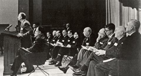 Black&amp;white image of man standing at podium with several more men seated on stage behind him.