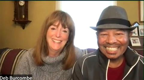A screenshot of Burcombe and Harris sitting together on a couch, both smiling