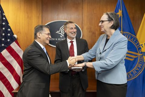 Rathmell holds handa on copy of the Constitution while shaking hands with HHS secretary, and her husband looks on, smiling.