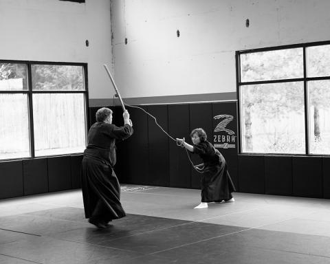 B&amp;W photo of man in martial arts pants and tunic holds up rod facing woman in similar uniform, slightly crouching, holding up rod and rope. 