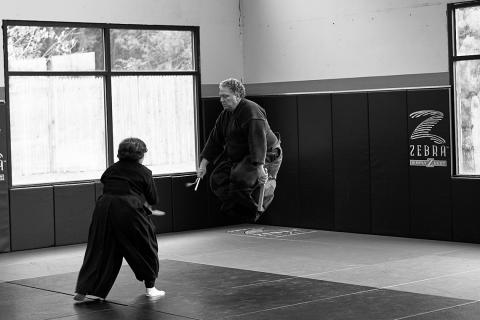 Black &amp; white photo of a woman in martial arts pants and tunic slightly crouches holding a sword as she faces a man, dressed similarly, in mid jump.