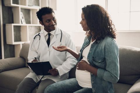 Pregnant woman seated on couch speaks to doctor sitting beside her.