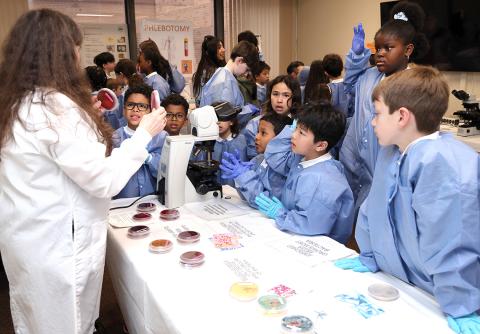 Group of kids, some looking in awe, others looking grossed out, stand around table as a tech holds up a petri dish of bacteria.