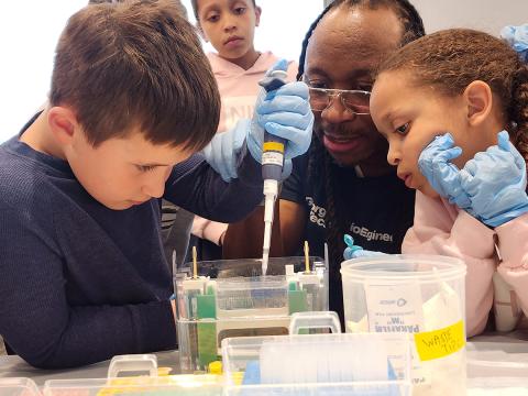 Dr. Platt guide's child's hand as he pipettes over a tray and other kids look on.