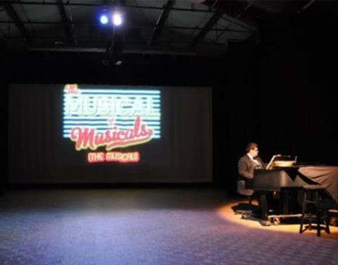 Tittermary sits at piano on stage, with Musical of Musicals on screen behind