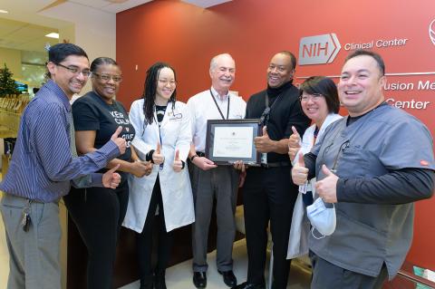 A group of 6 people giving a thumbs up gesture, framing an older man holding a framed certificate